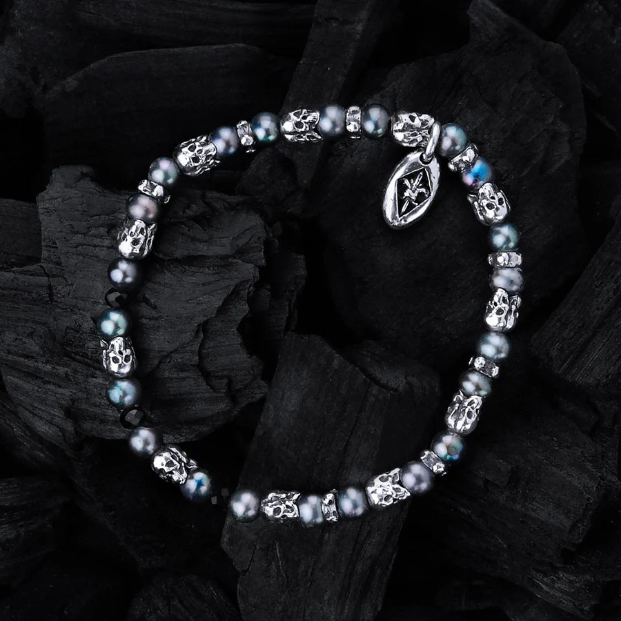 Produktfotografie des Elcraft Armbands mit sich abwechselnden schwarzen Perlen und silbernen Triple-Skull-Elementen im Totenkopf-Design während das Band auf schwarzer Kohle liegt