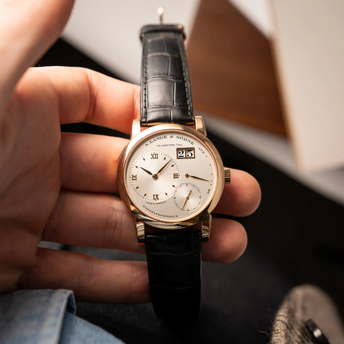 Mann hält die A. Lange & Söhne Lange 1 der Referenz 101.032 mit 38,5mm-Roségold-Gehäuse und dem für das Modell typische, asymmetrisch angeordnete Zifferblatt mit Großdatum in seiner Hand und präsentiert die Uhr in die Kamera