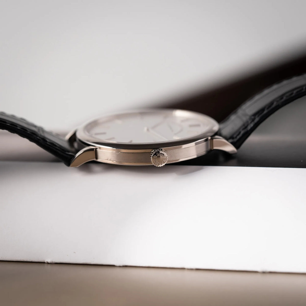 Produktfotografie der A.Lange Söhne Saxonia Thin der Referenz 211.026 im Weißgold-Gehäuse mit weiß-silbernem Zifferblatt am schwarzen Lederarmband zeigt die seitliche Gehäuseflanke und die Aufzugskrone