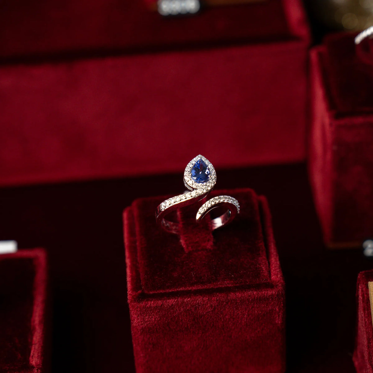 Produktfotografie des weißgoldenen Damenrings ABTN-155-1 von Lunatica Roma mit einer offenen, diamantbesetzten Ringschiene und einem Tansanit-Tropfen als Highlight, während der RIng auf einer weinroten Juweliers-Deko präsentiert wird