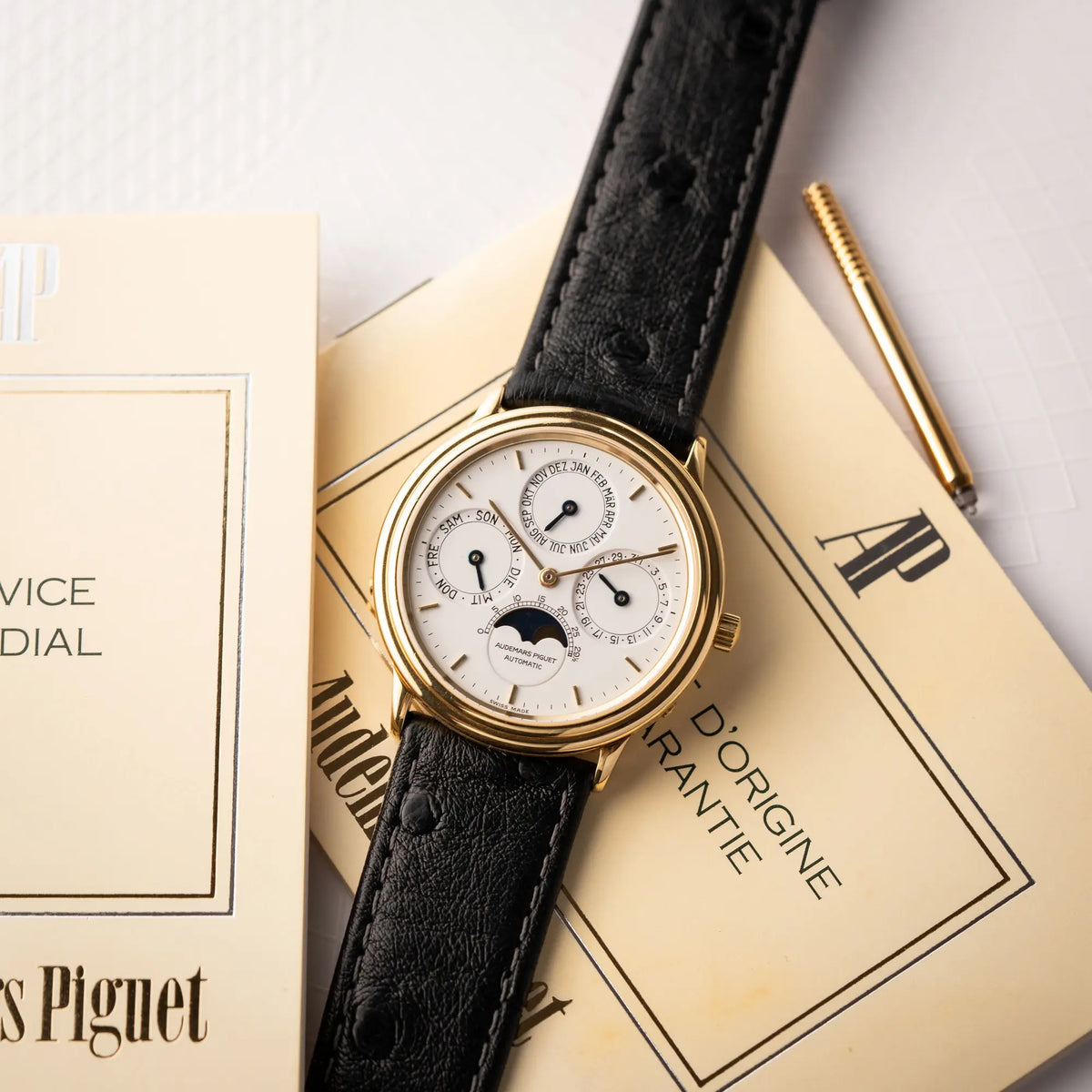 Produktfotografie der Audemars Piguet Quantième Perpétual der Referenz 5548BA mit 36mm-Gelbgold-Gehäuse und weißem Zifferblatt an einem schwarzen Lederband zusammen mit Stellstift