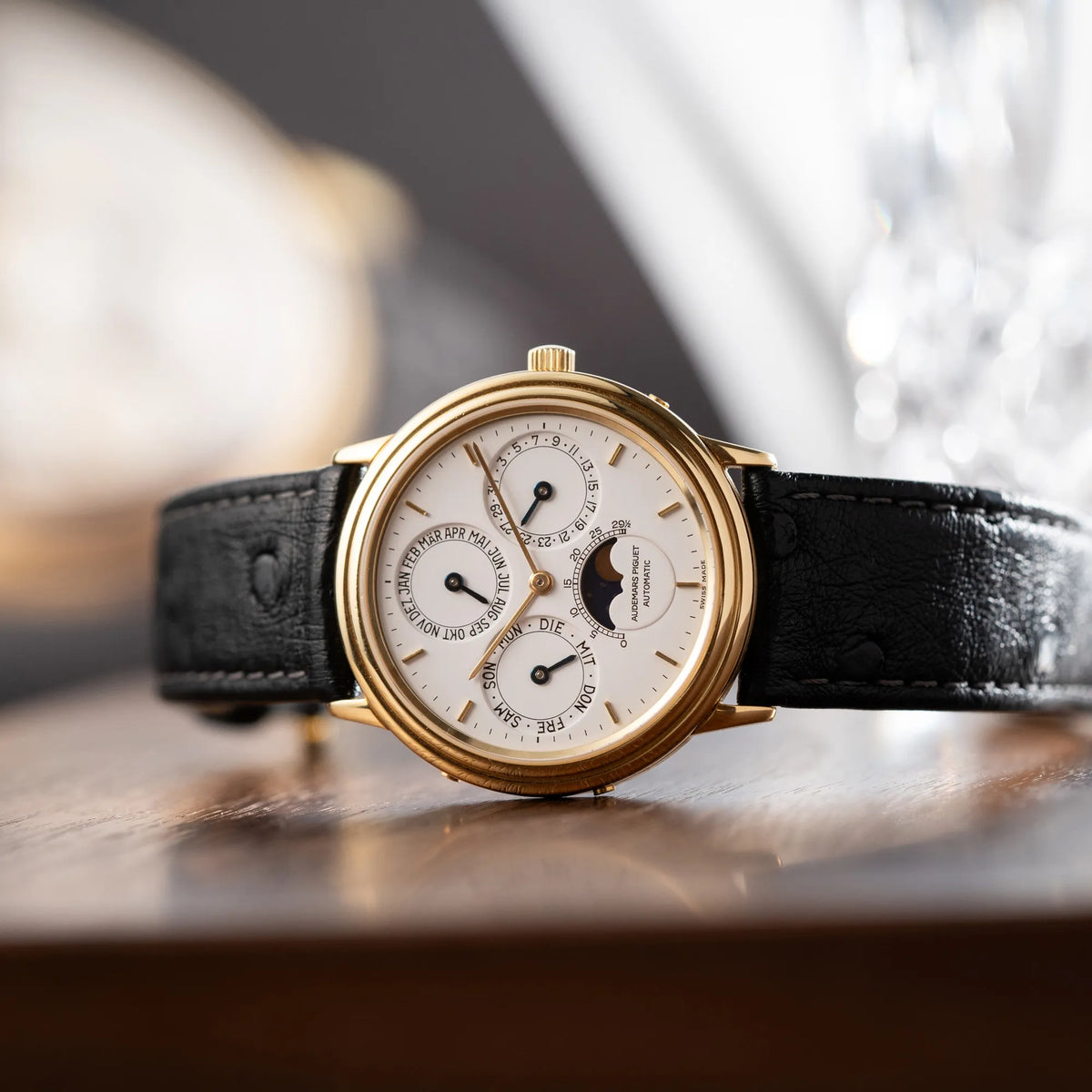 Produktfotografie der seitlich auf dem Uhrengehäuse liegenden Audemars Piguet Quantième Perpétual der Referenz 5548BA mit 36mm-Gelbgold-Gehäuse und weißem Zifferblatt an einem schwarzen Lederband