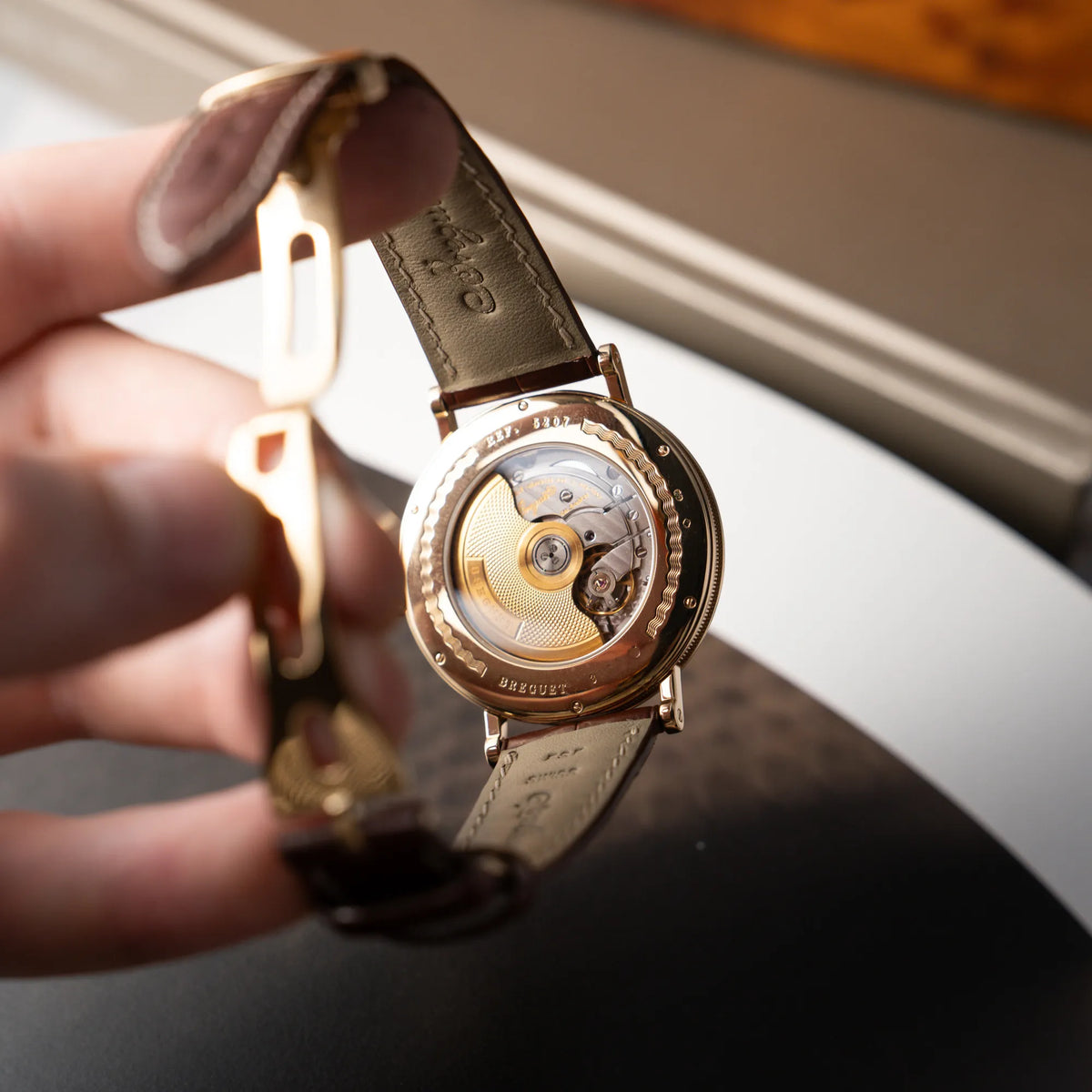 Mann hält die gelbgoldene Breguet Armbanduhr des Models Classic Retrograde 5207 am braunen Lederband in der Hand und präsentiert den Gehäuseboden mit SIchtglas