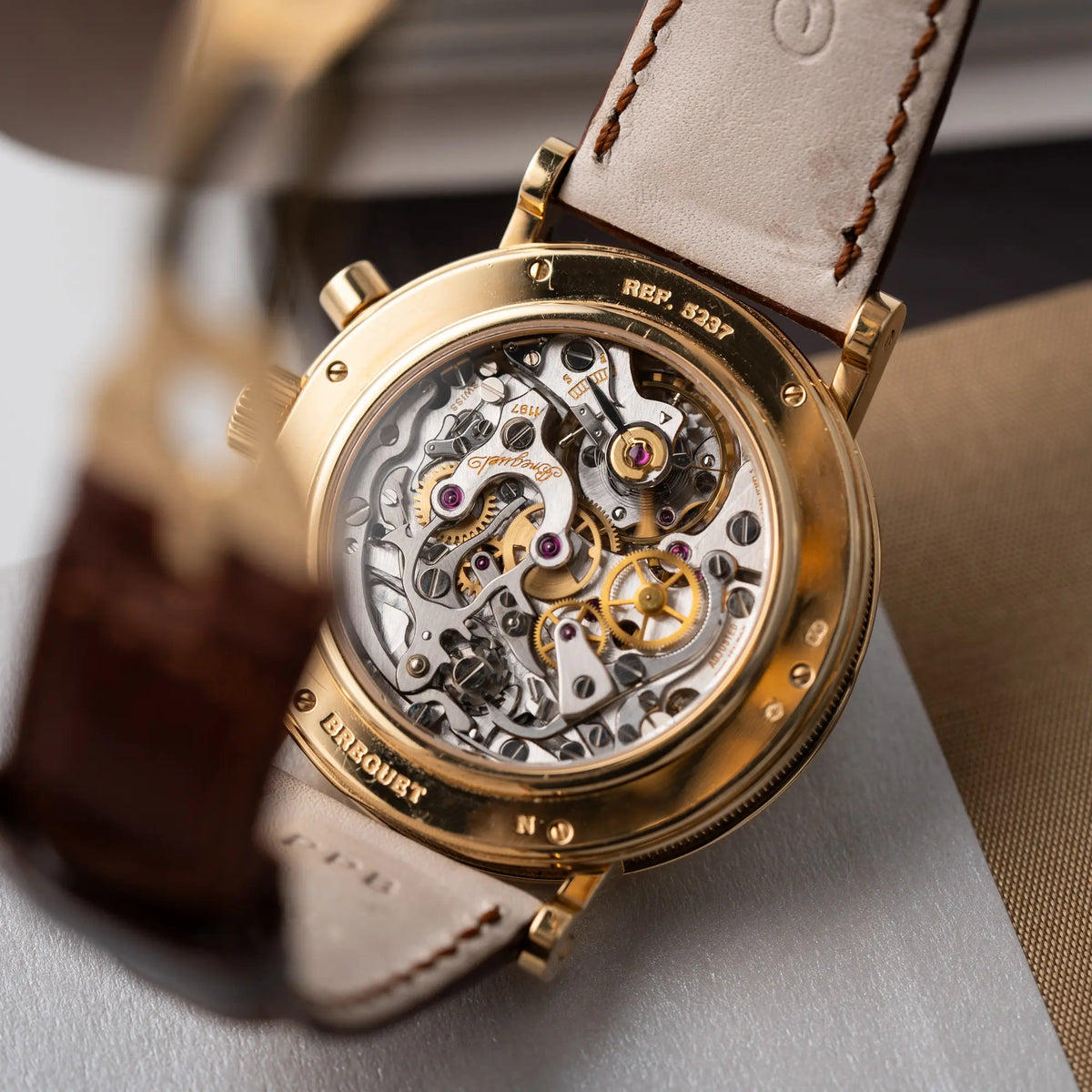 Produktfotografie zeigt das Handaufzugs-Kaliber durch den Gehäuseboden mit Sichtglas von dem Breguet Classique Chronograph der Referenz 5237 in Gelbgold mit silbernem guillochiertem Zifferblatt am braunem Lederband