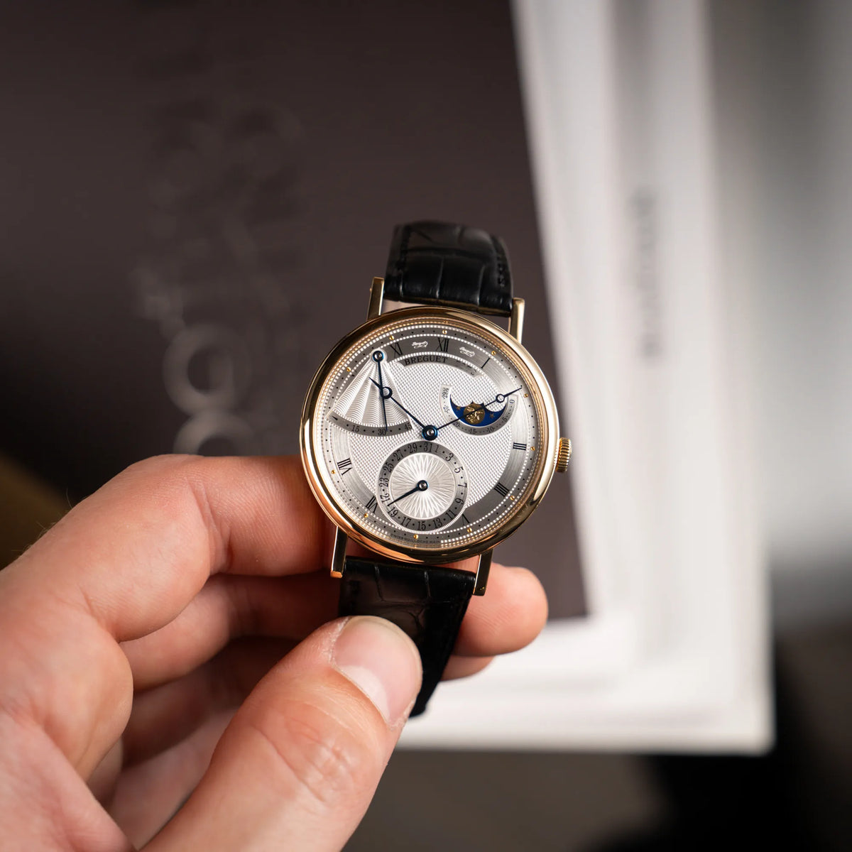 Mann hält die Breguet Classique Mondphase 7137 in Gelbgold mit silbernen, verschieden guillochiertem Zifferblatt und präsentiert die Uhr in die Kamera