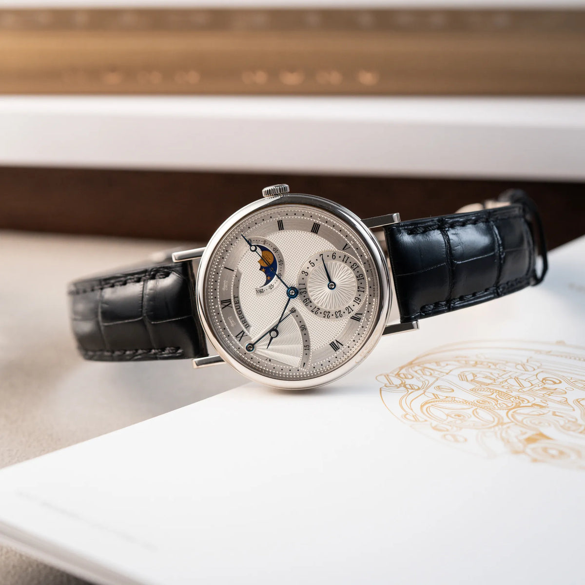 Produktfotografie der seitlich auf dem Gehäuse liegenden Breguet Classique Moonphase der Referenz 7137 in Weissgold mit 3-fach guillochiertem Zifferblatt und Mondphase-Anzeige