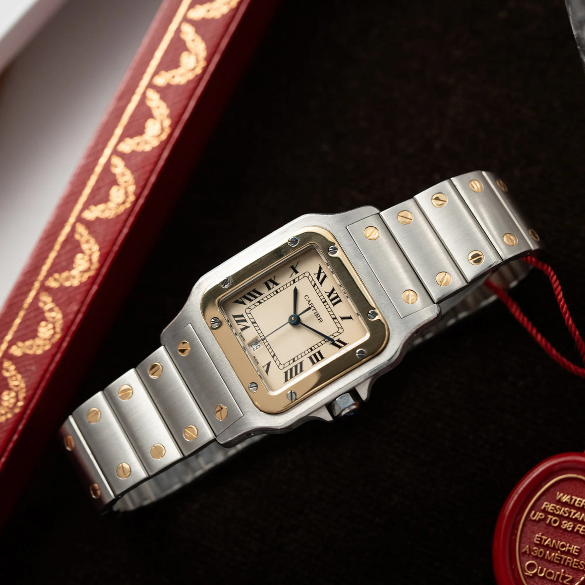Produktfotografie derCartier Santos Galbee BiColor der Referenz 187901 mit Stahl-Gold-Gehäuse am Stahlband mit dem Cartier-typischen Schrauben, die ebenfalls in Gelbgold gehalten sind, und einem cremfarbenen Zifferblatt mit klassischen römischen Indizes auf einem Cartier-Juwelierstablet