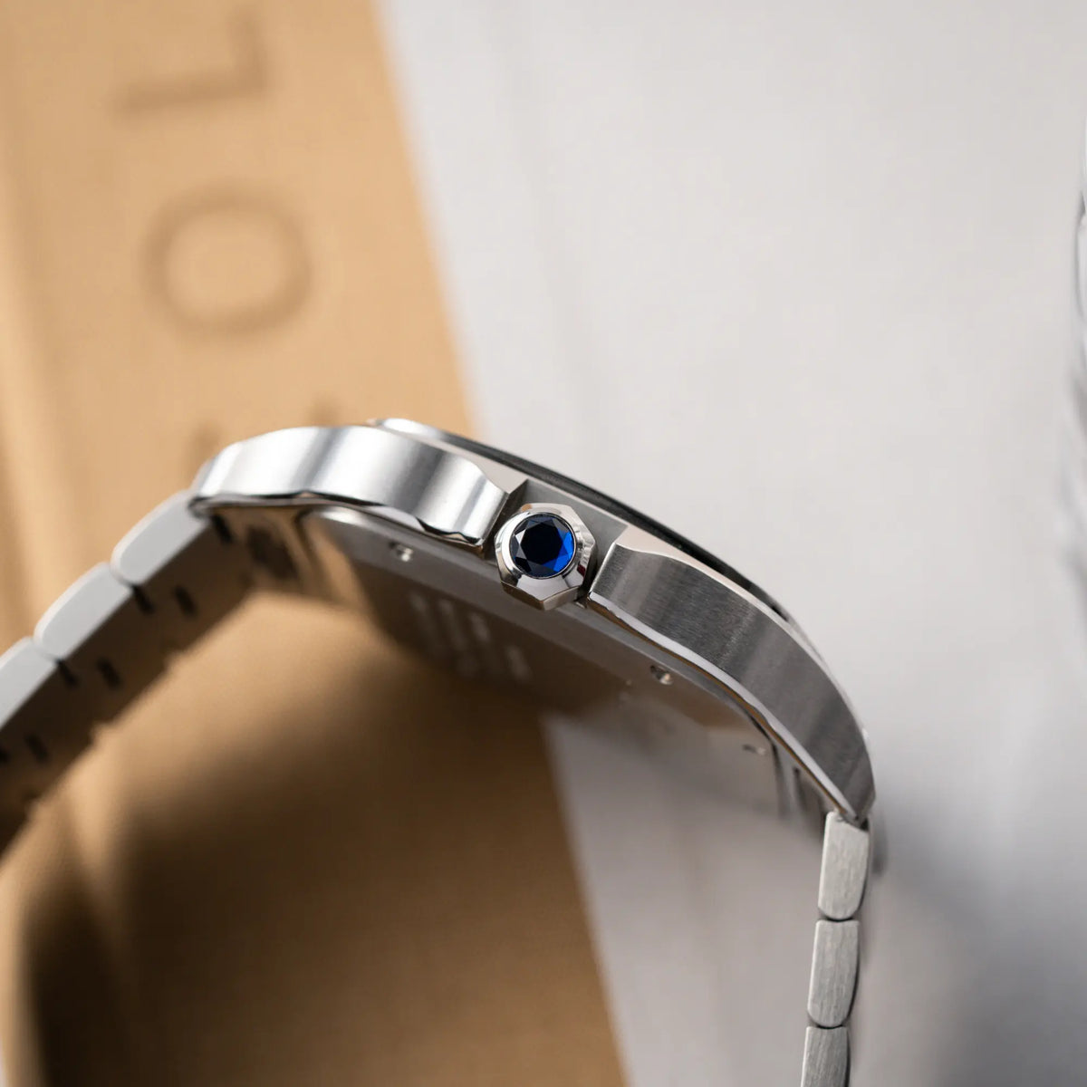 Produktfotografie der Cartier Santos de Cartier als großes Modell der Referenz WSA0018 mit dem klassischen weißen Zifferblatt und römischen Indizes zeigt die Aufzugskrone mit Cartier typischen blauen Edelstein und die verschiedenen Phasen des Bandanstoßes mit polierten und satinierten Oberflächen