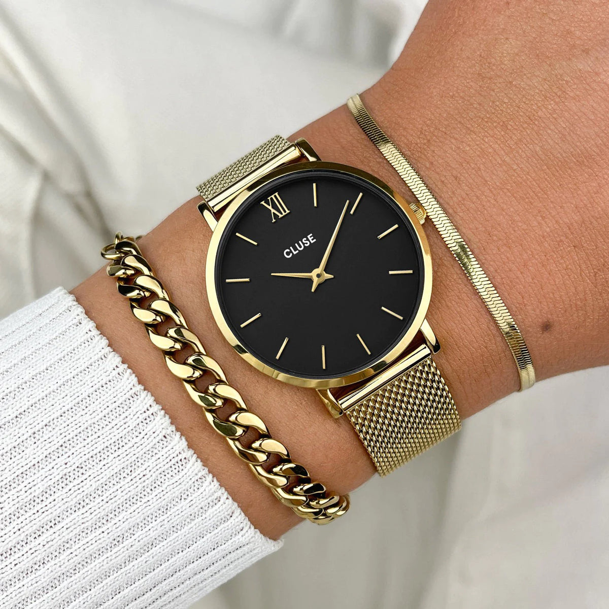 Frau trägt Minuit Mesh Uhr mit goldfarbenen Gehäuse am Handgelenk und kombiniert diese mit goldenen Schmuckstücken