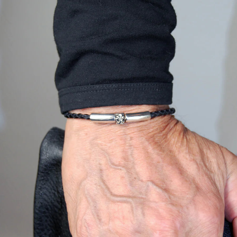 Mann trägt ein feines, schwarzes, geflochtenes Lederarmband mit glattem Silberverschluss, welcher durch ein Lilien-Detail verziert wurde, am Handgelenk