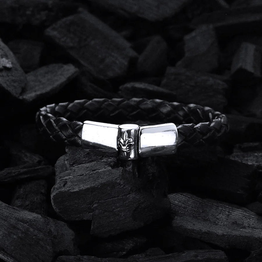 Produktfoto des schwarzen, geflochtenen Lederarmbands mit glattem Silberverschluss und einer zentralen Lilie als Design-Highlight des Verschlusses vor schwarzer Holzkohle