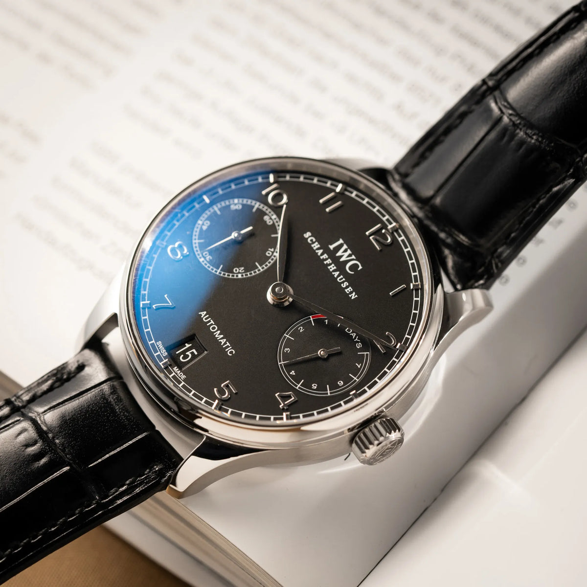 Produktfotografie der flach auf einem Buch liegenden IWC Portugieser 7 Days IW500109 mit schwarzem Zifferblatt am schwarzen Lederband
