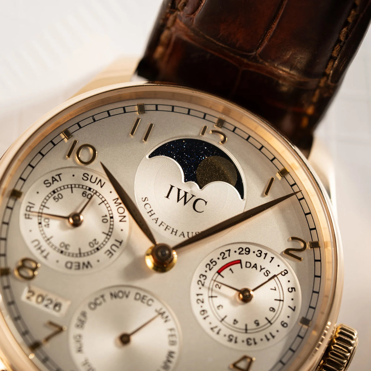 Detailaufnahme des Portugieser Ewiger Kalender IW502213 von IWC in einem 42mm-Rosegold-Gehäuse am braunem IWC Lederband mit einem sanften weißem Zifferblatt mit Fokus auf der Mondphase-Anzeige bei 12 Uhr