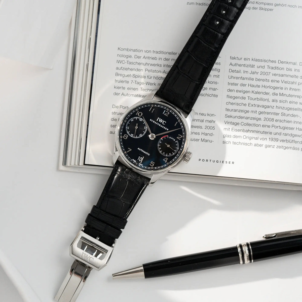Produktfotografie der Portugieser 7 Days Automatic IW500109 Uhr von IWC Schaffhausen am schwarzen Lederband mit schwarzem Zifferblatt neben einem schwarz-silbernen Kugelschreiber