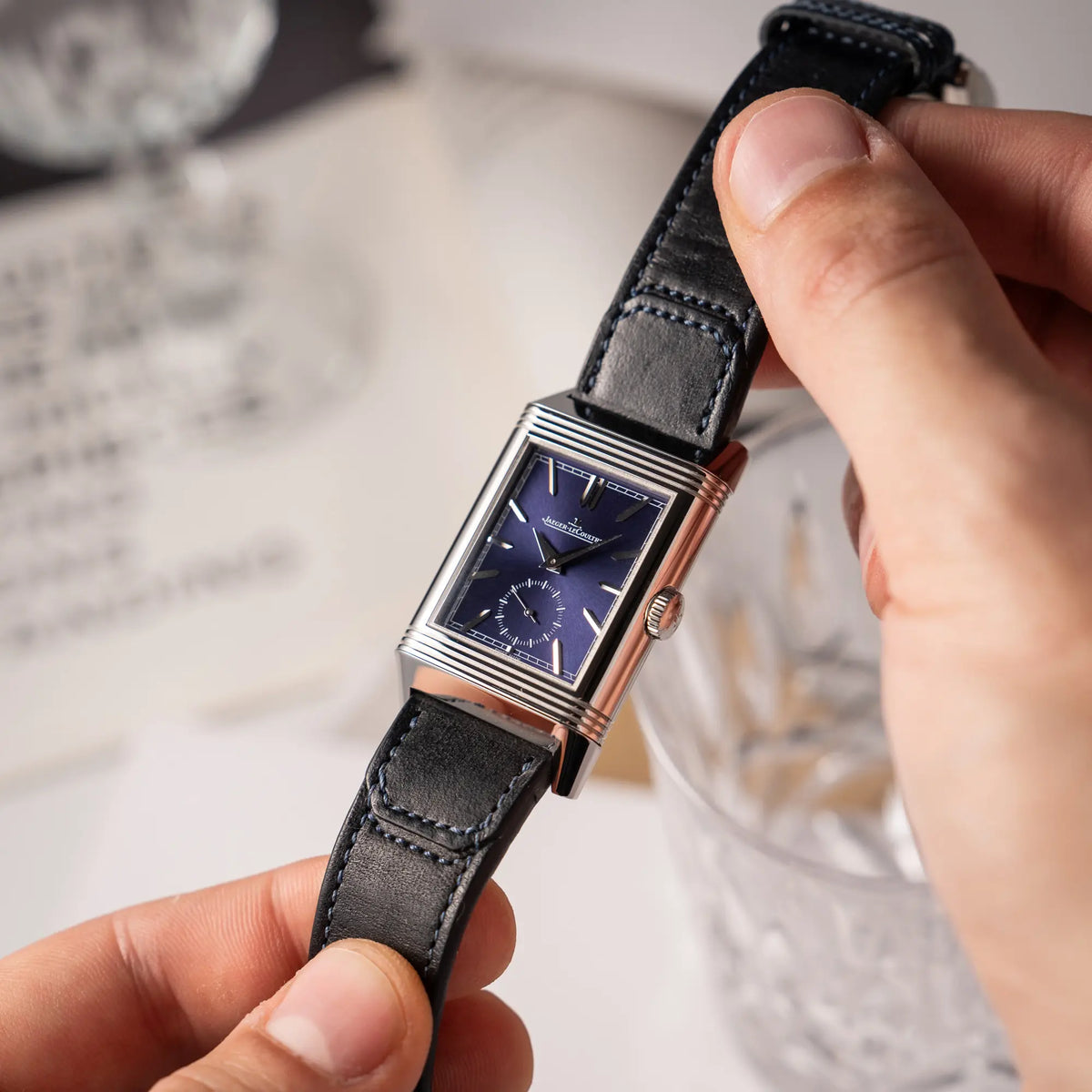 Mann hält die Jaeger-LeCoultre Reverso Duoface Q3988482 mit blauem Zifferblatt und weißem Night-and-Day-Zifferblatt am schwarzen Lederband in de Fingern und präsentiert die Uhr in die Kamera