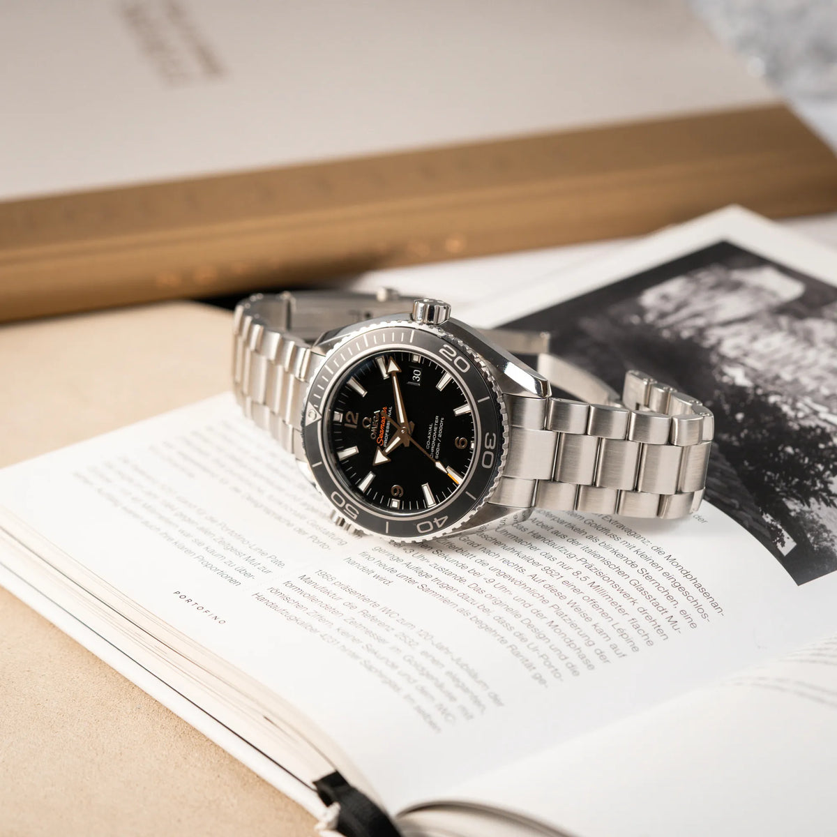 Produktfotografie der Omega Seamaster Planet Ocean 232.30.46.21.01.001 mit 45mm Gehäuse und schwarzem Zifferblatt mit orangen Details, während die Uhr auf dem aufgeschlagenen Uhrenbuch liegt
