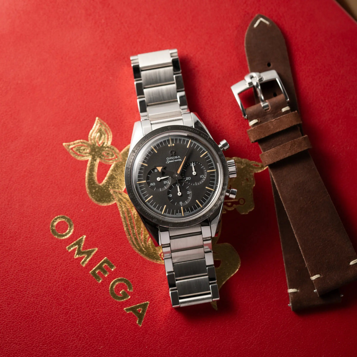 Produktfotografie der Omega Speedmaster 1957 Trilogy Limited Edition mit einem anthrazitfarbenen Zifferblatt mit Vintage-Look und dem original Omega-Stahlband im Ed-White-Design zusammen mit dem zusätzlichen braunem Omega-Lederband, während die Uhr auf der roten Omega-Box liegt