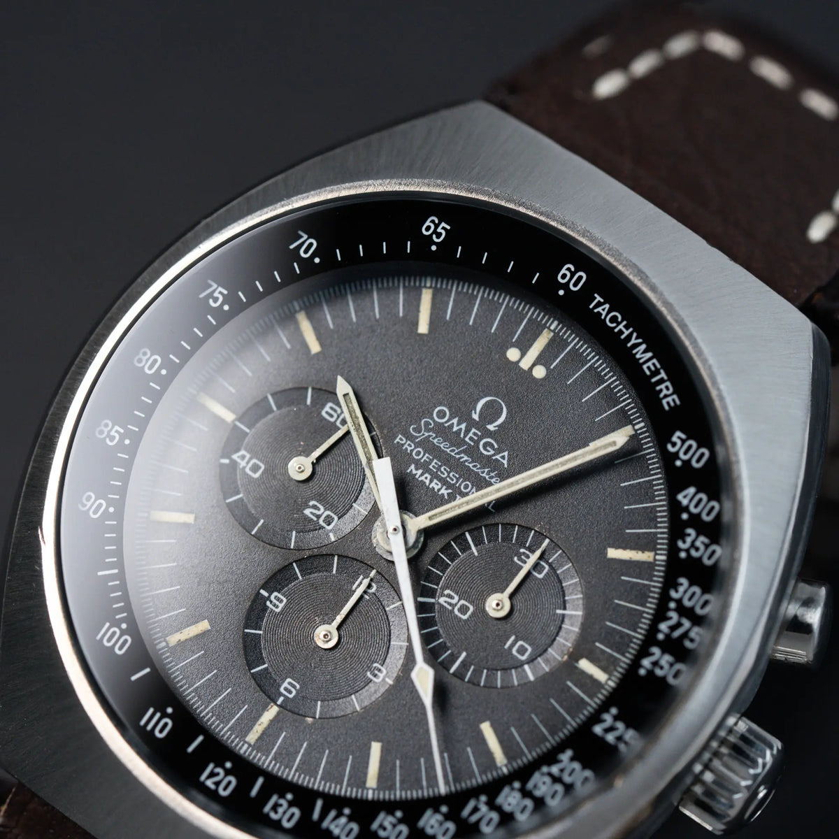 Detailaufnahme des schwarzen Zifferblatts und der Lünette/Gehäuse der Omega Speedmaster Moonwatch Mark 2 an einem braunen Lederarmband