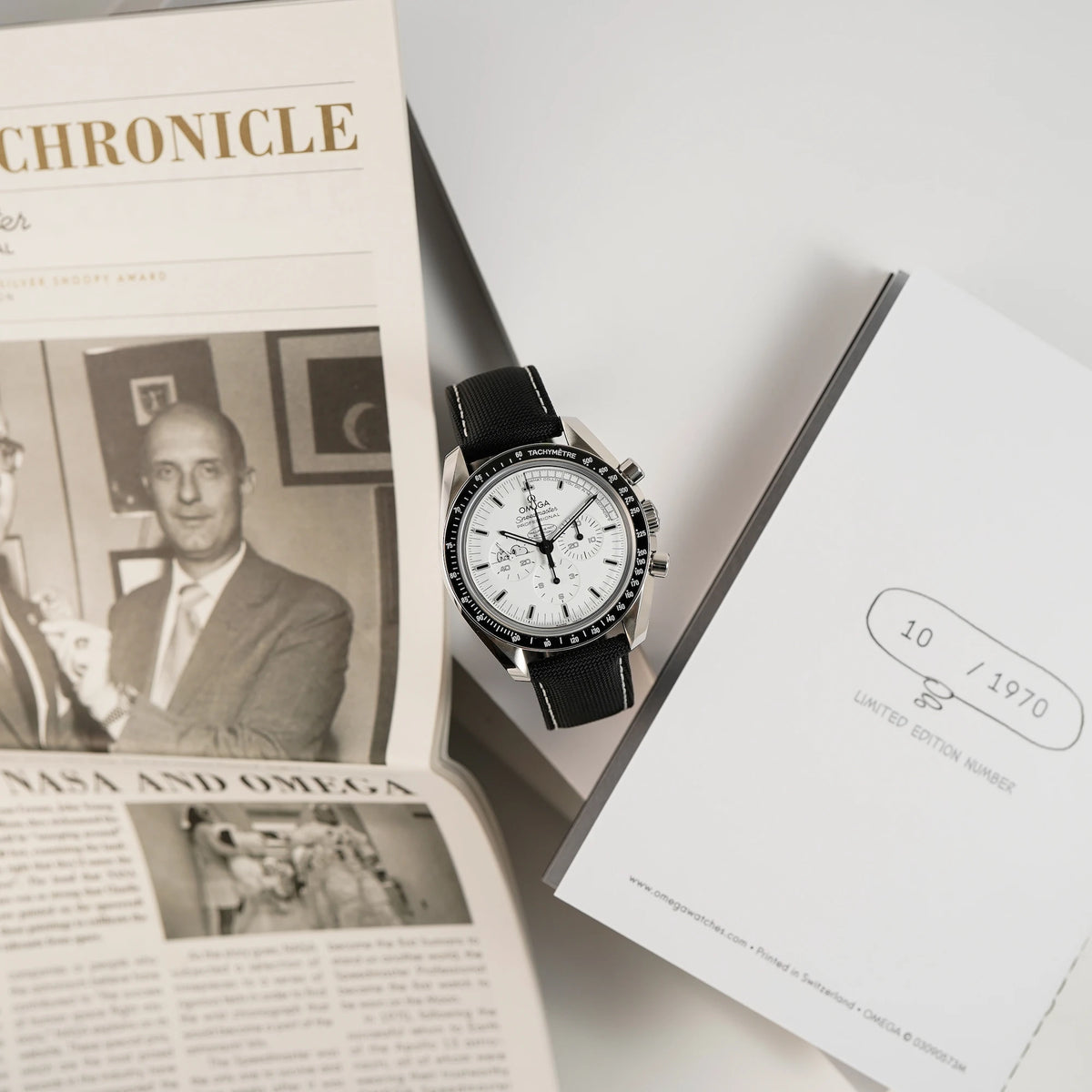 Produktfotografie der limitierten Omega Speedmaster Moonwatch Silver Snoopy mit dem weißen Zifferblatt zusammen mit dem "Apollo 13 Chronicle" Heft aus dem Lieferumfang