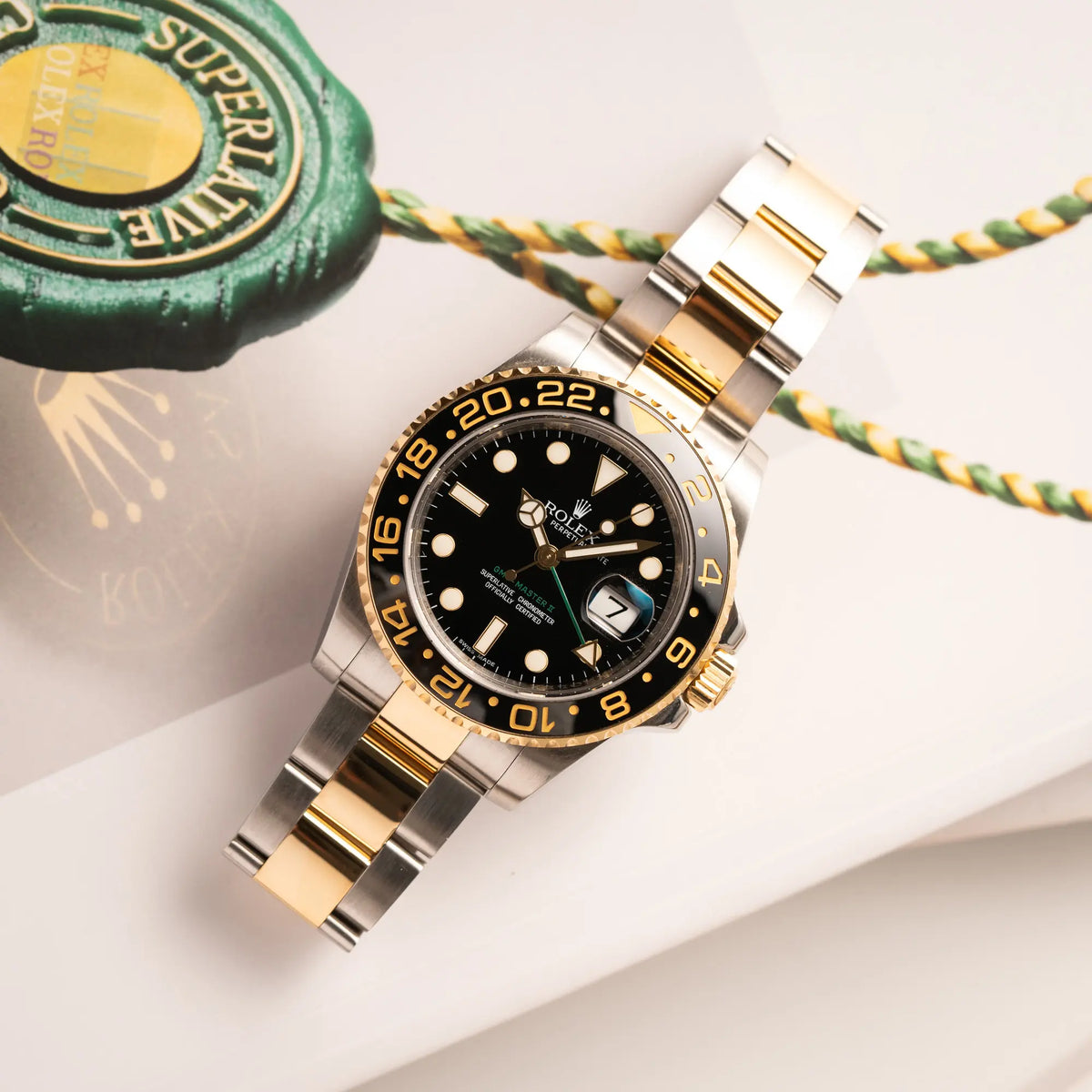 Produktfotografie der Rolex GMT-Master II der Referenz 116713LN in Stahl-Gold mit schwarzem Zifferblatt und dem grünen GMT-Zeiger, während die Uhr flach auf einem Buch liegt, welches das Rolex Hangtag zeigt