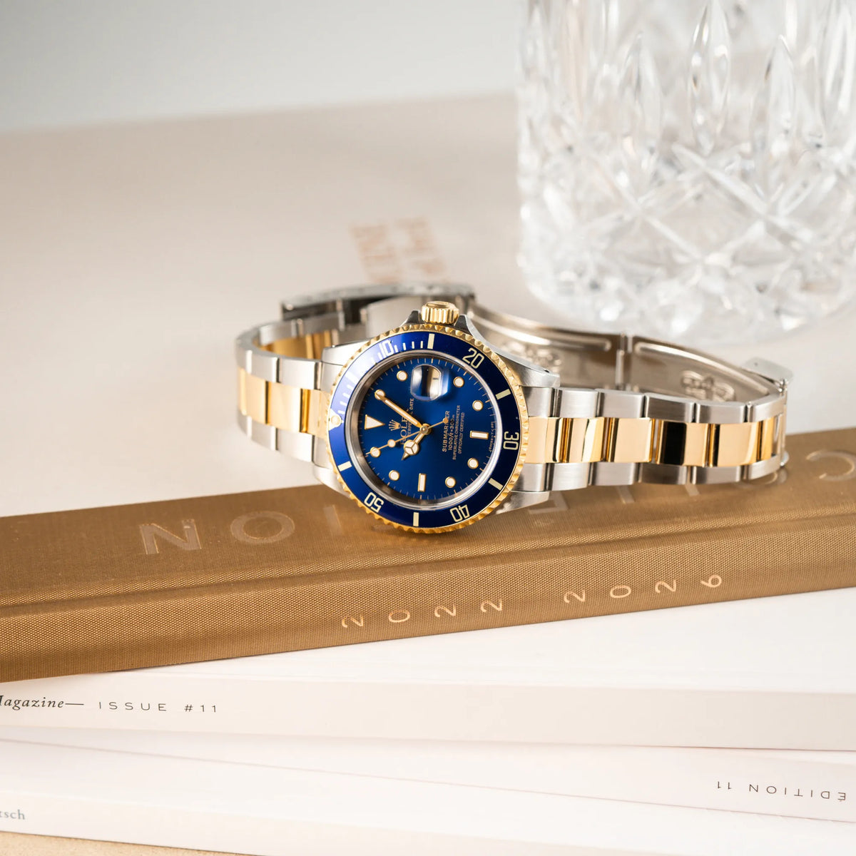 Produktfotografie der der bicolor Rolex Submariner Date der Referenz 16613 mit dem Spitznamen "Bluesy" in Stahl-Gold mit blauem Zifferblatt während die Uhr auf einem Uhrenbuch liegt