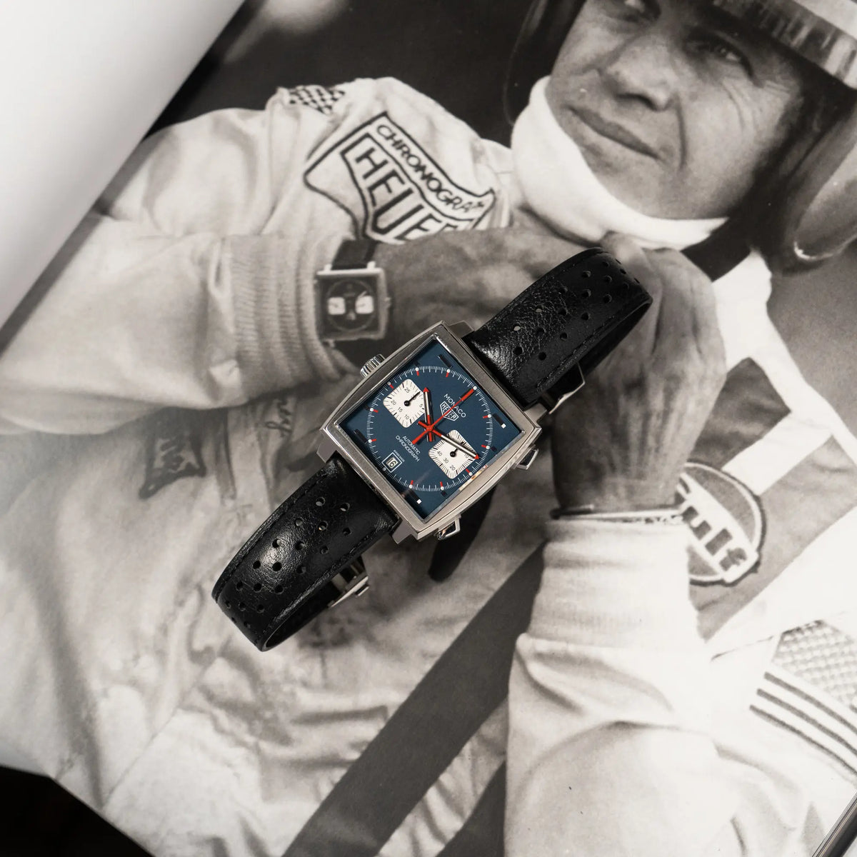 Produktfotografie des Tag Heuer Monaco Calibre 11 Chronographen CAW211P mit dem klassischen blauen Zifferblatt und roten Details auf Zeigern und Indizes mit schwarz-weißem Steve McQueen Foto als Hintergrund