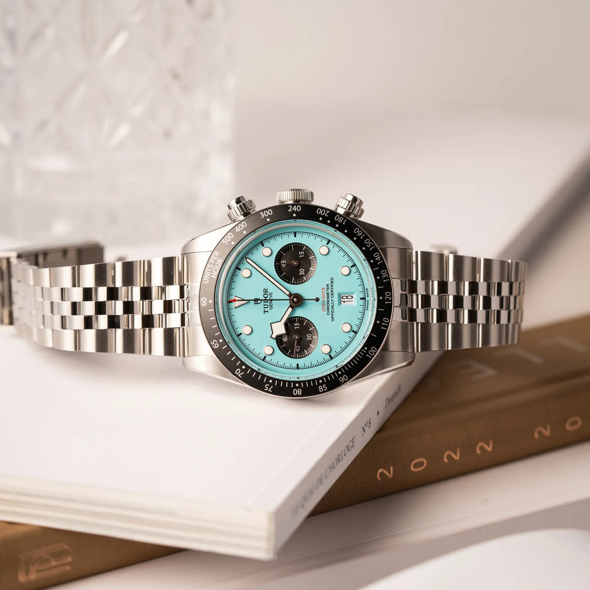 Tudor Black Bay Chronograph Flamingo mit dem türkis-blauen Zifferblatt mit schwarzen Totalisatoren und schwarzer Lünette mit Zähler-Skala liegt seitlich auf einem Buch über Uhren
