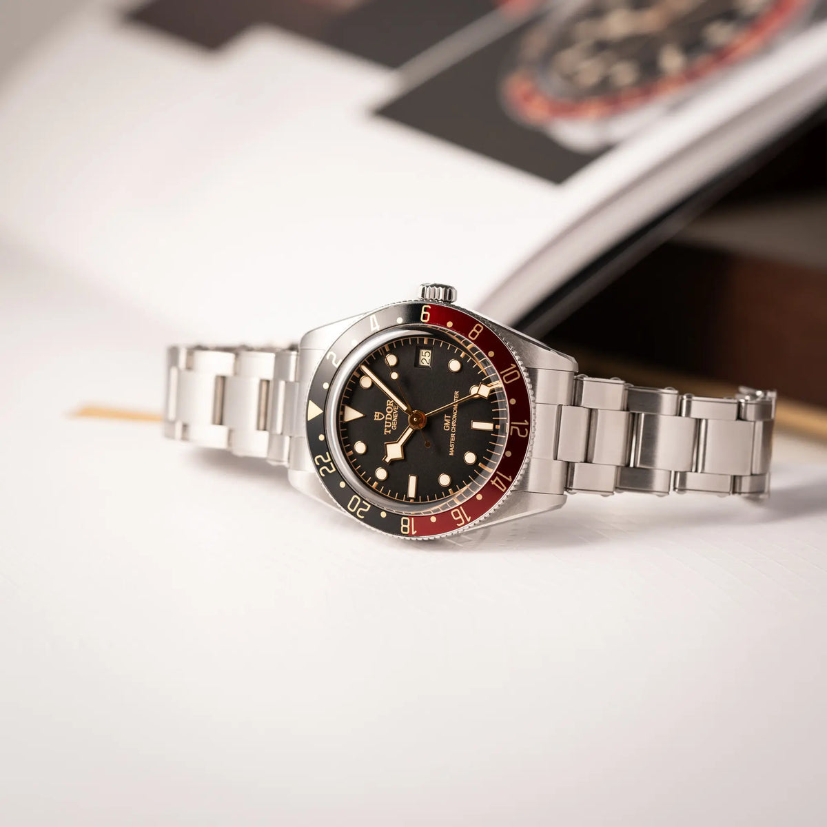 Tudor BlackBay GMT Coke der Referenz 7939G1A0NRU mit der schwarz-roten Lünette und bronzefarbenen Details am Edelstahlarmband liegt seitlich auf dem Uhrengehäuse auf einem Uhrenbuch