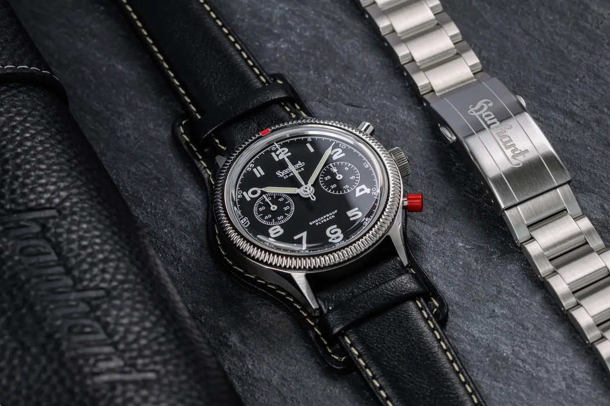 Produktfotografie der Hanhart 417 ES 1954 Heritage Flyback mit 39mm-Gehäuse und schwarzem Zifferblatt am schwarzen Lederband, während das Edelstahlarmband daneben liegt