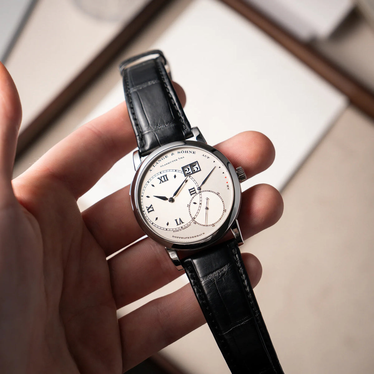 Mann hält die Großen Lange 1 mit Platingehäuse und silberfarbenem Zifferblatt am schwarzen Lederband von A. Lange & Söhne in seiner Hand und präsentiert die Uhr