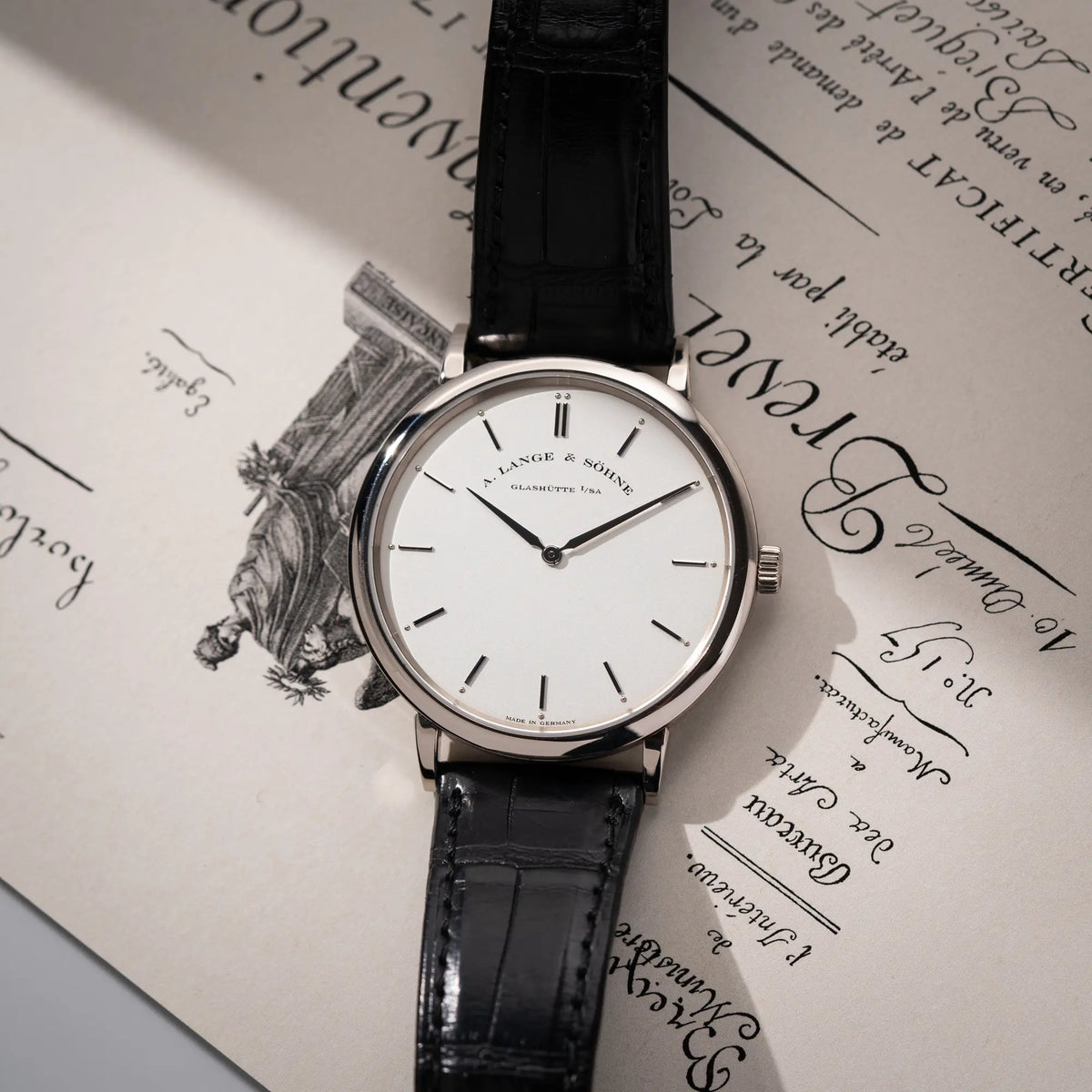 Produktfotografie zeigt die flach liegende A.Lange Söhne Saxonia Thin der Referenz 211.026 im Weißgold-Gehäuse mit weiß-silbernem Zifferblatt am schwarzen Lederarmband