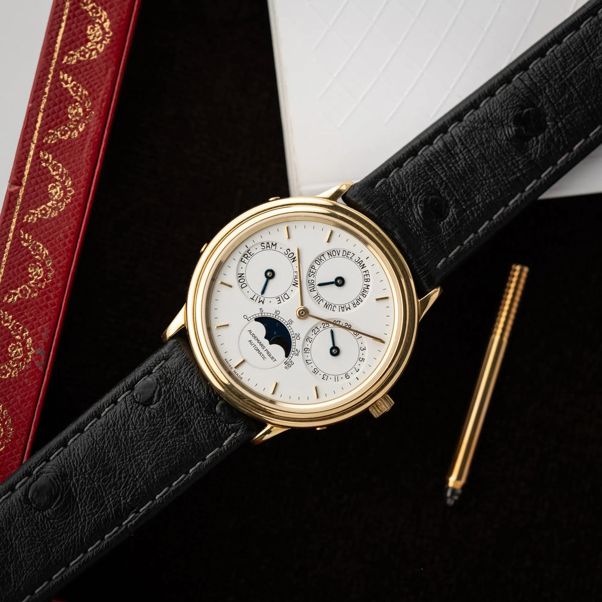 Fotografie der Audemars Piguet Quantième Perpétual der Referenz 5548BA mit 36mm-Gelbgold-Gehäuse und weißem Zifferblatt an einem schwarzen Lederband zusammen mit dem gelbgoldenen Stellschift
