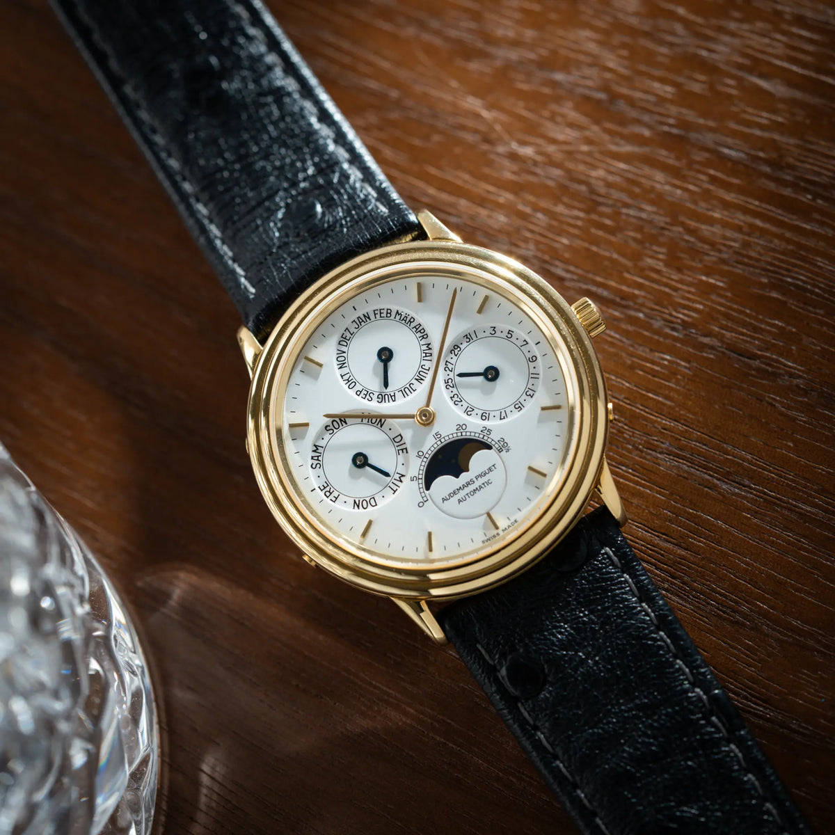 Audemars Piguet Quantième Perpétual der Referenz 5548BA mit 36mm-Gelbgold-Gehäuse und weißem Zifferblatt an einem schwarzen Lederband liegt flach auf einem Holztisch