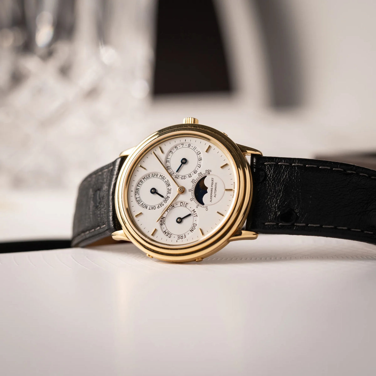Produktfotografie der seitlich auf dem Uhrengehäuse liegende Audemars Piguet Quantième Perpétual der Referenz 5548BA mit 36mm-Gelbgold-Gehäuse und weißem Zifferblatt an einem schwarzen Lederband