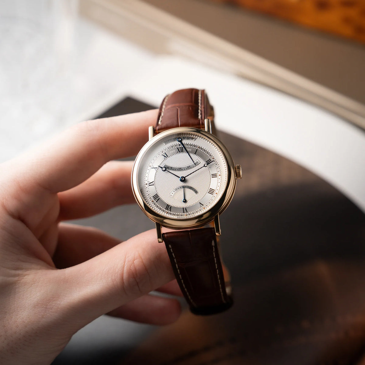 Mann hält die gelbgoldene Breguet Armbanduhr des Models Classic Retrograde 5207 am braunen Lederband in der Hand und präsentiert die Uhr der Kamera