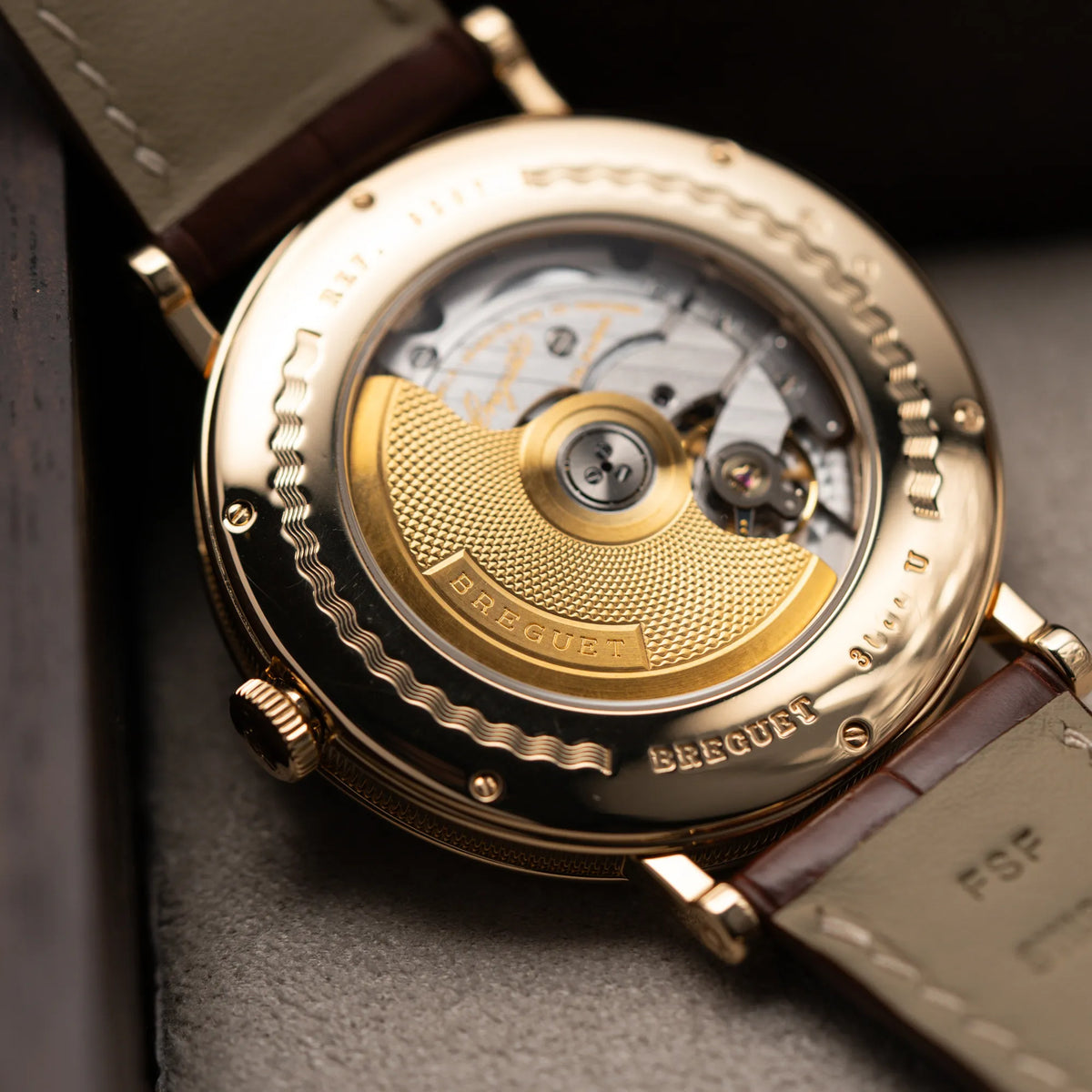 Detailaufnahme des Gehäusebodens der gelbgoldenen Breguet Armbanduhr des Models Classic Retrograde 5207 am braunen Lederband