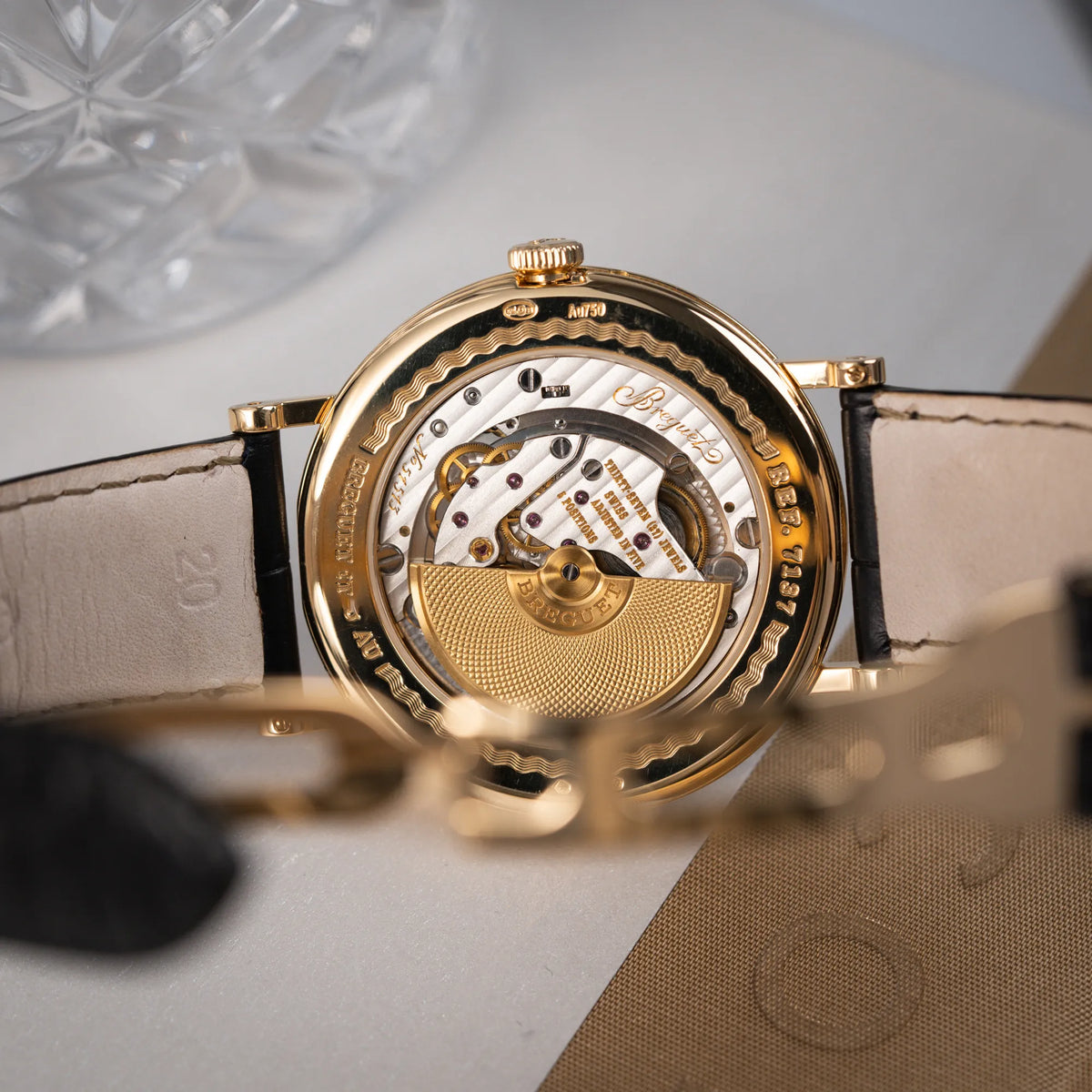Produktfotografie zeigt das Uhrwerk der Breguet Classique 7137BA mit einem Gelbgoldgehäuse in klassischer Form mit einem silbernen Zifferblatt mit jeweils einem anderen handgefertigten Guilloche-Muster für jede Komplikation  durch den gläsernen Gehäuseboden