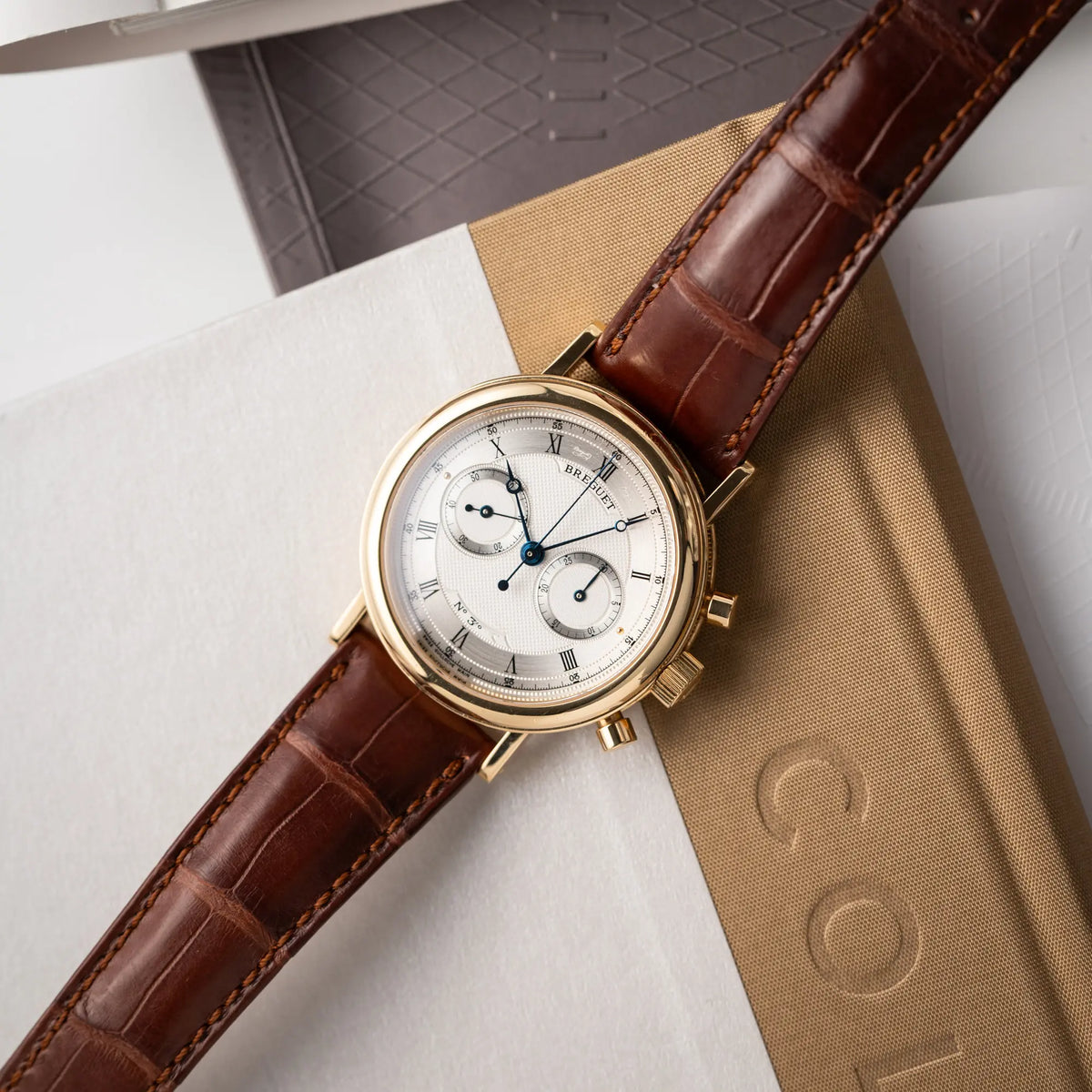 Breguet Classique Chronograph der Referenz 5237 in Gelbgold mit silbernem guillochiertem Zifferblatt am braunem Lederband  liegt flach auf einem Uhrenbuch