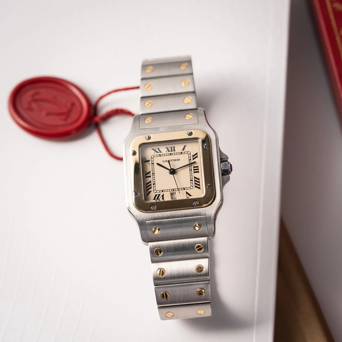 Produktfotografie der Cartier Santos Galbee BiColor der Referenz 187901 mit Stahl-Gold-Gehäuse am Stahlband  mit dem Cartier-typischen Schrauben, die ebenfalls in Gelbgold gehalten sind, und einem cremfarbenen Zifferblatt mit klassischen römischen Indizes zusammen mit dem roten Cartier-Hangtag