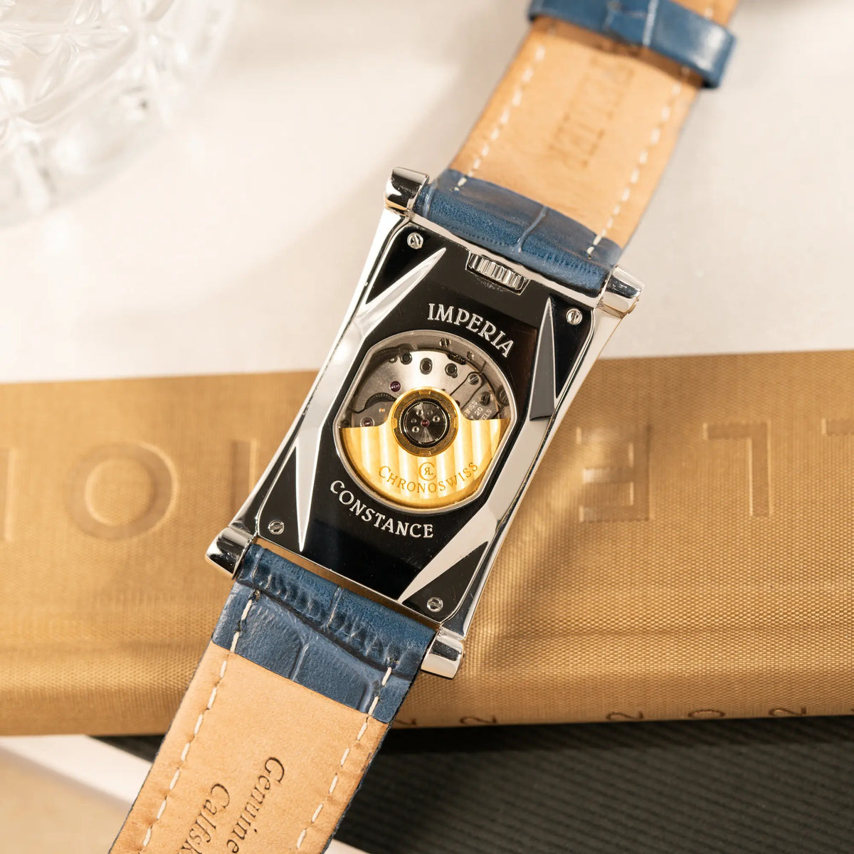 Produktfotografie der Chronoswiss Imperia Constanze mit diamantbesetzten-Stahlgehäuse mit schwarzem Zifferblatt an einem petrol-blauem Lederband zeigt den Gehäuseboden mit Sichtglas für das Uhrwerk
