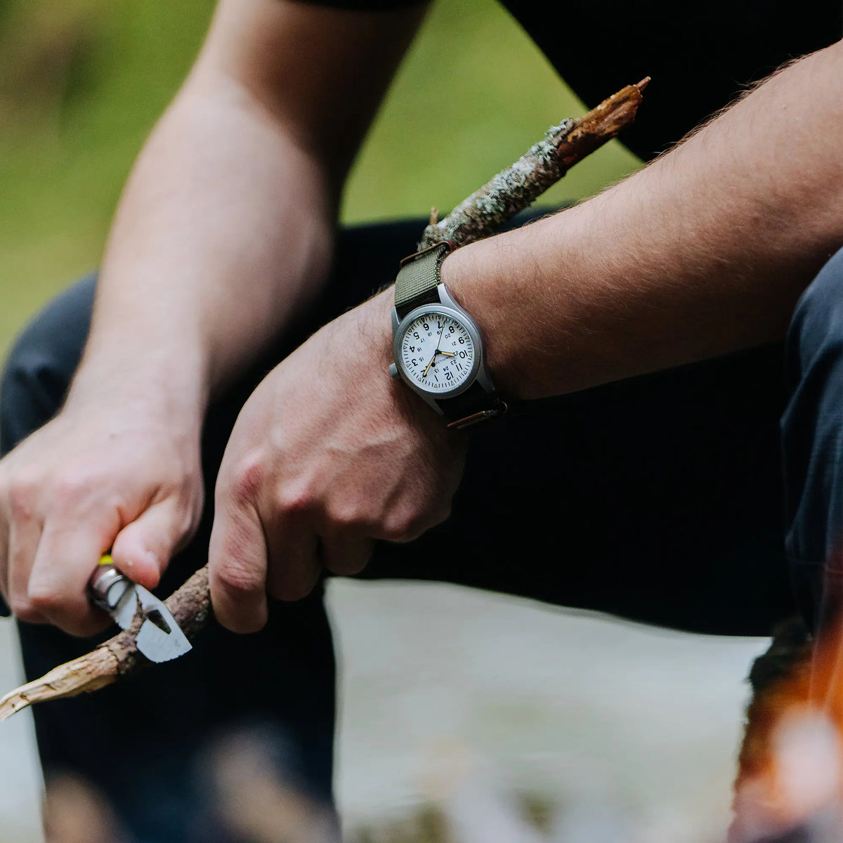 Mann trägt die Hamilton Khaki Field Mechanical H69439411 mit weißem Zifferblatt am olivgrünen Nato-Armband an seinem Handgelenk, während dieser im Wald einen Stock schnitzt