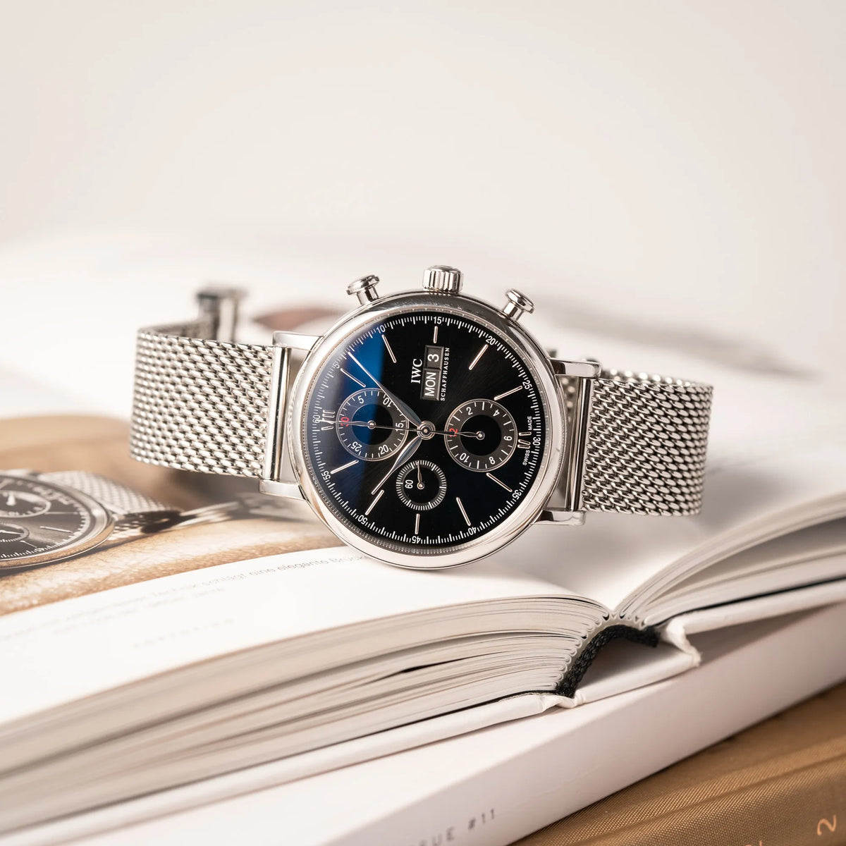 IWC Portofino Chronograph der Referenz IW391002 mit schwarzem Zifferblatt an einem Mesh-Armband liegt auf einem Buch, das die Uhr abzeigt