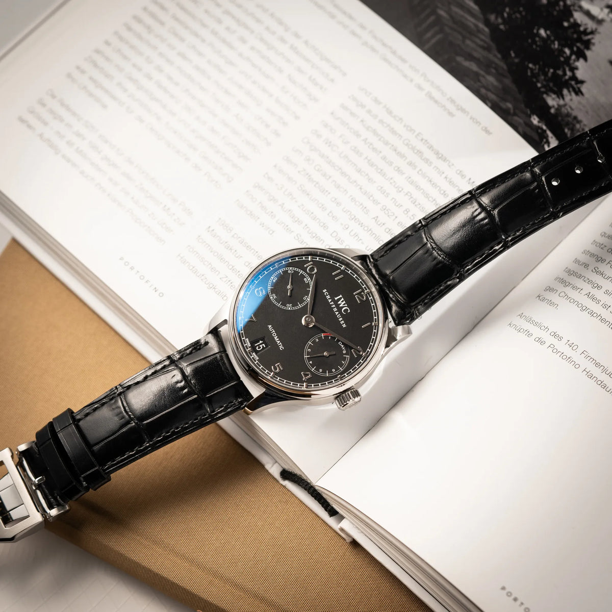 IWC Portugieser 7 Days IW500109 mit schwarzem Zifferblatt am schwarzen Lederband liegt auf einem Buch über Uhren
