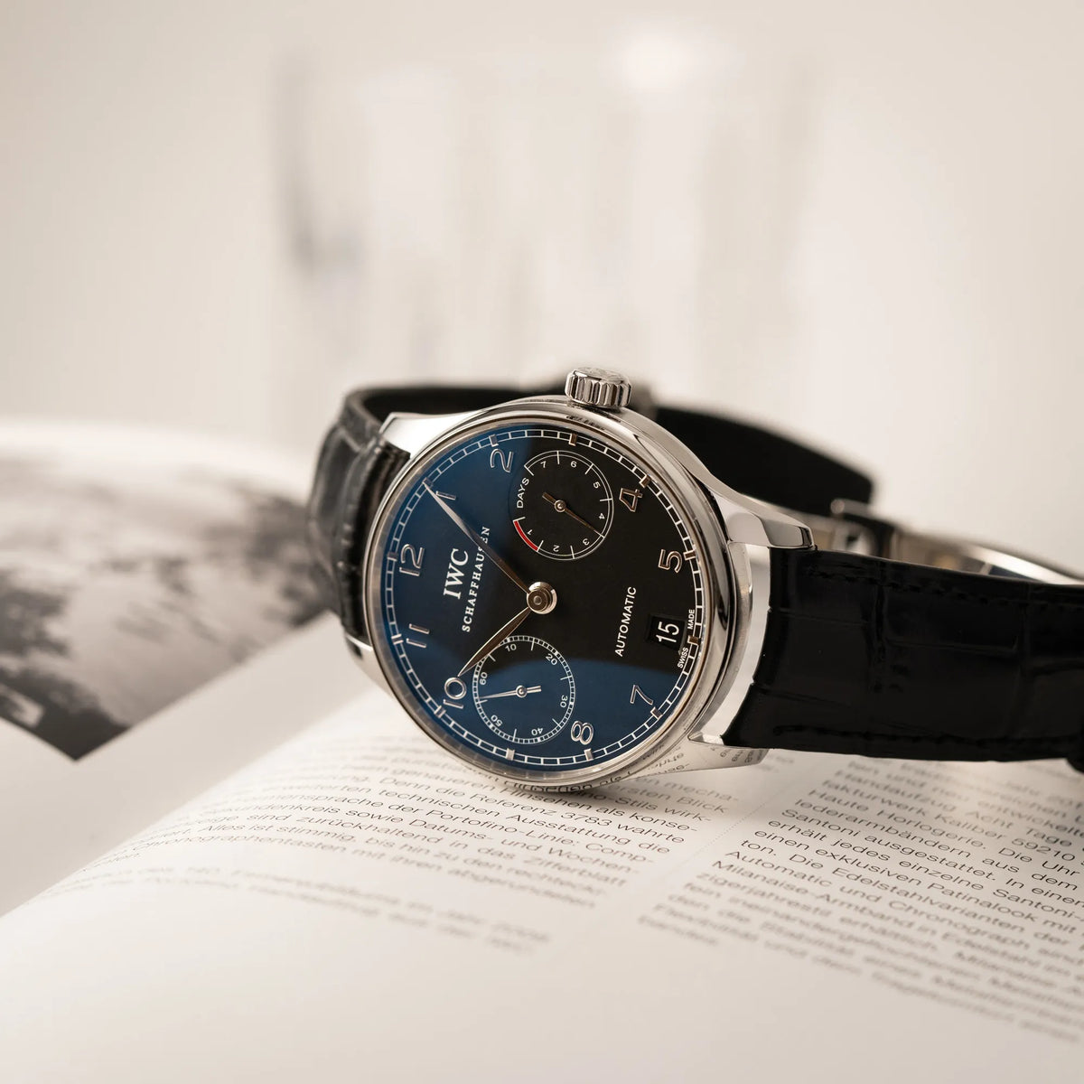 IWC Portugieser 7 Days IW500109 mit schwarzem Zifferblatt am schwarzen Lederband liegt auf der seitlichen Gehäuseflanke auf einem Buch
