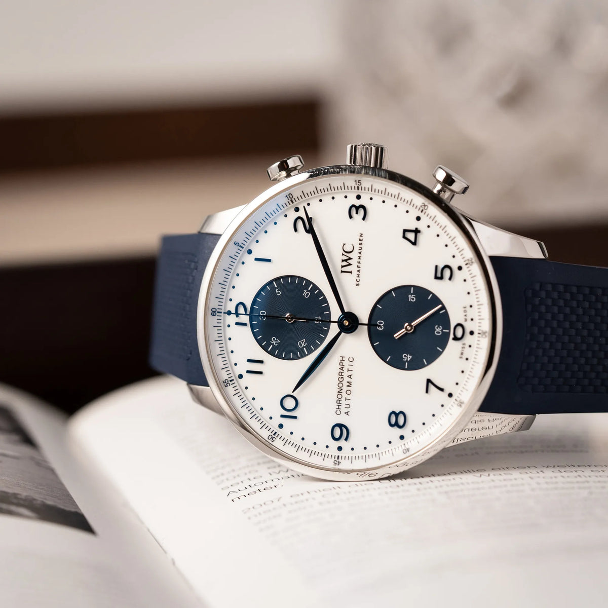 Detailreiche Produktfotografie der seitlich auf dem Uhrengehäuse liegenden IWC Portugieser Chronograph 41 der Referenz IW371620 mit weißem Zifferblatt und dunkelblauen Totalisatoren am passenden dunkelblauen Kautschukarmband