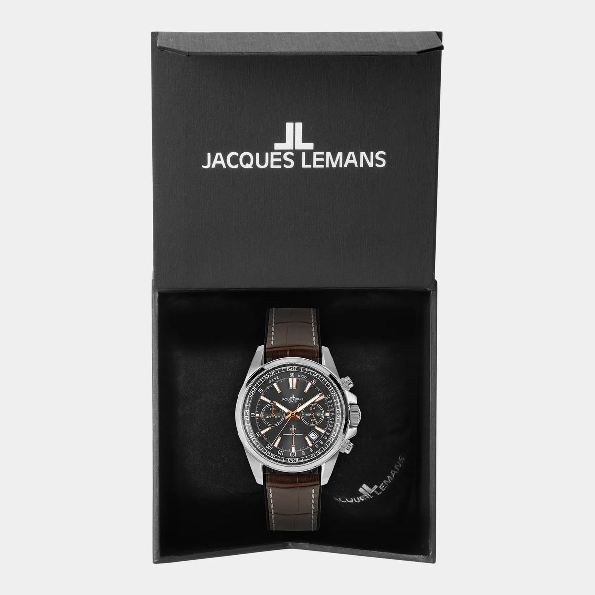Uhrenbox mit der Jacques Lemans 1-2117W Chronograph-Armbanduhr für Herren mit einem anthrazitfarbenen Zifferblatt, roségoldfarbenen Ziegern und Edelstahl-Gehäuse an einem braunem Lederarmband mit einer groben Struktur und mit weißer Ziernaht 
