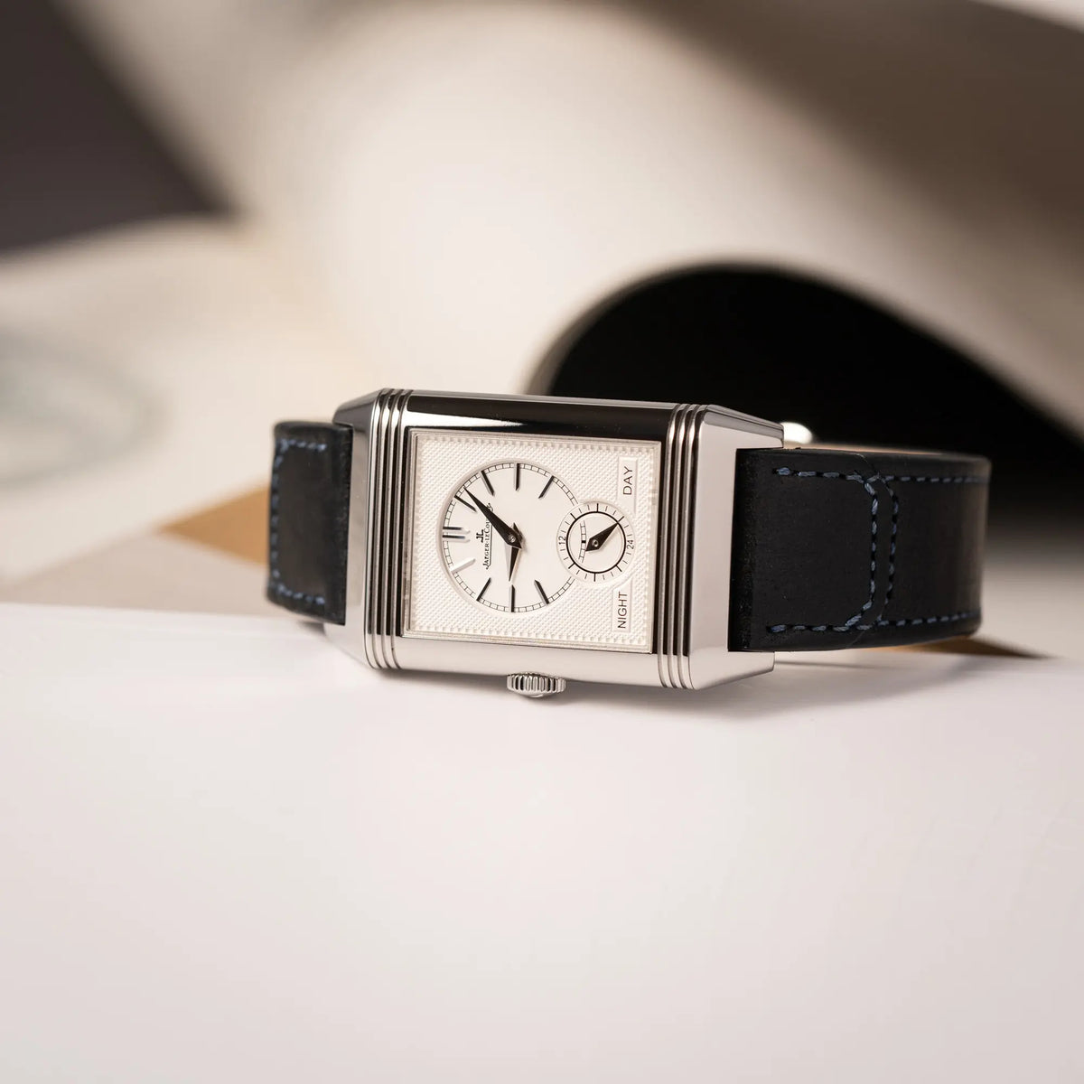 Produktfotografie der seitlich auf dem Uhrengehäuse leigenden Jaeger-LeCoultre Reverso Duoface Q3988482 mit blauem Zifferblatt und weißem Night-and-Day-Zifferblatt am schwarzen Lederband
