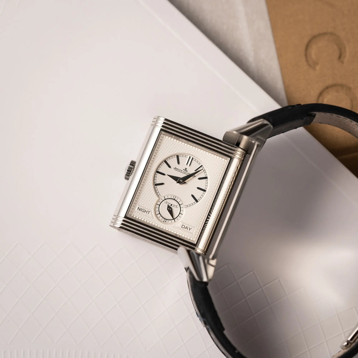 Produktfotografie der seitlich auf dem Uhrengehäuse liegenden Jaeger-LeCoultre Reverso Duoface Q3988482 mit blauem Zifferblatt und weißem Night-and-Day-Zifferblatt am schwarzen Lederband mit betätigtem Reverso Mechanismus, zeigt das weiße Zifferblatt