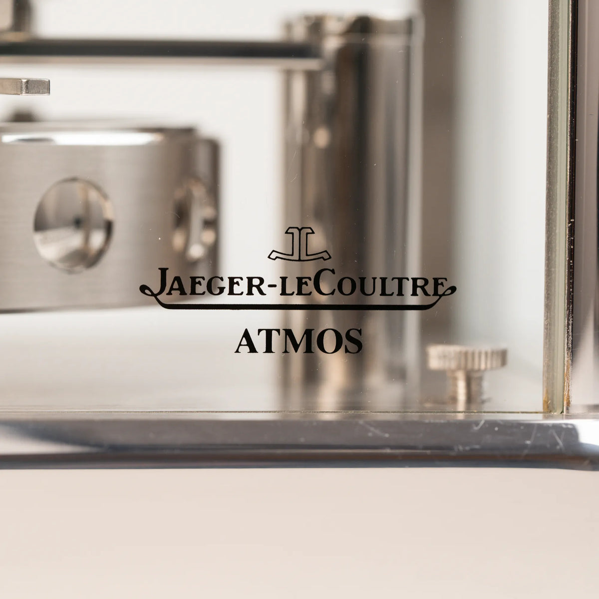 Detailaufnahme des "Jaeger-LeCoultre Atmos" Schriftzugs auf der vorderen Glastür der  Jaeger-LeCoultre Atmos Mondphase Tischuhr in rhodiniertem Stahl mit weißem Zifferblatt und blauen römischen Indizes