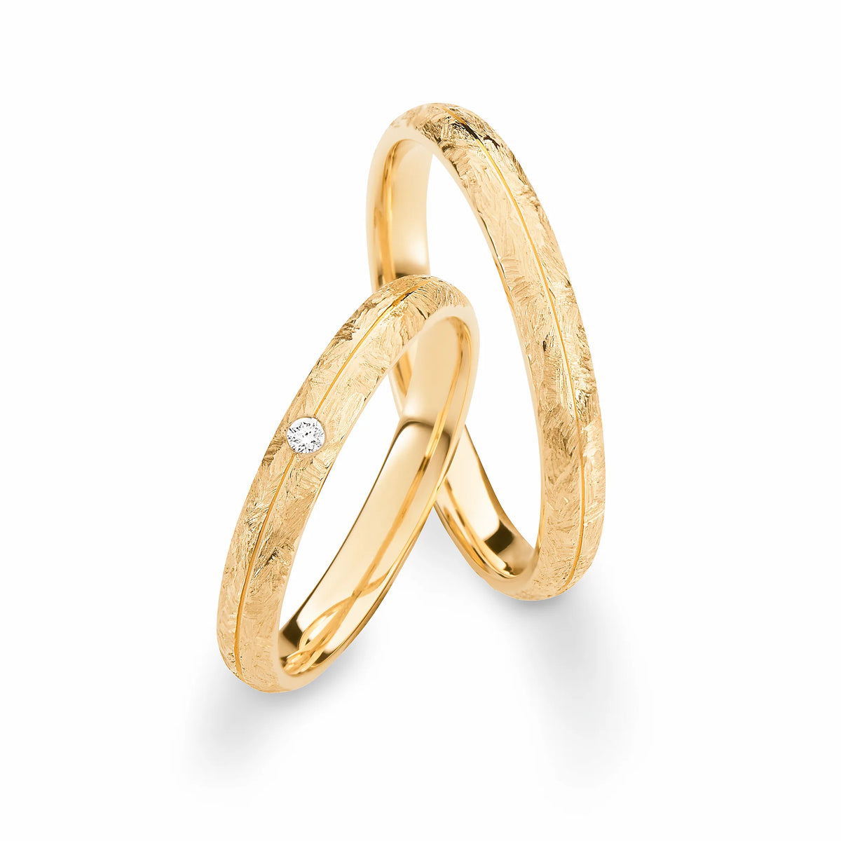 Produktfotografie des gelbgoldenen Max Kemper Trauringpaars mit wild gebürsteter Oberflächenstruktur und einer zentralen Nut, die sich um den Ring herumzieht, sowie einem zentralen Diamanten als Highlight des Damenrings